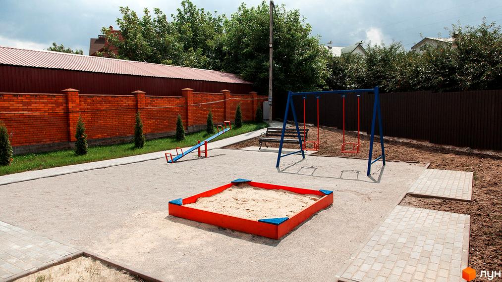 Дитячий майданчик у Котеджному містечку Затишне з пісочницею, гойдалкою та гойдалкою-балансиром. Навколо майданчика розташовані зелені насадження та цегляний паркан.