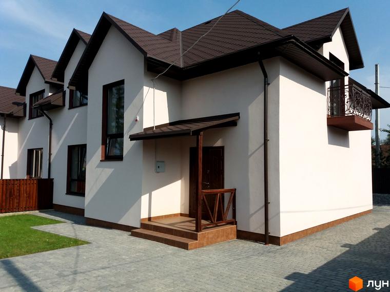 Зовнішній вигляд двоповерхового будинку з коричневим дахом та білими стінами. Навколо будинку розташована бруківка та зелена галявина.