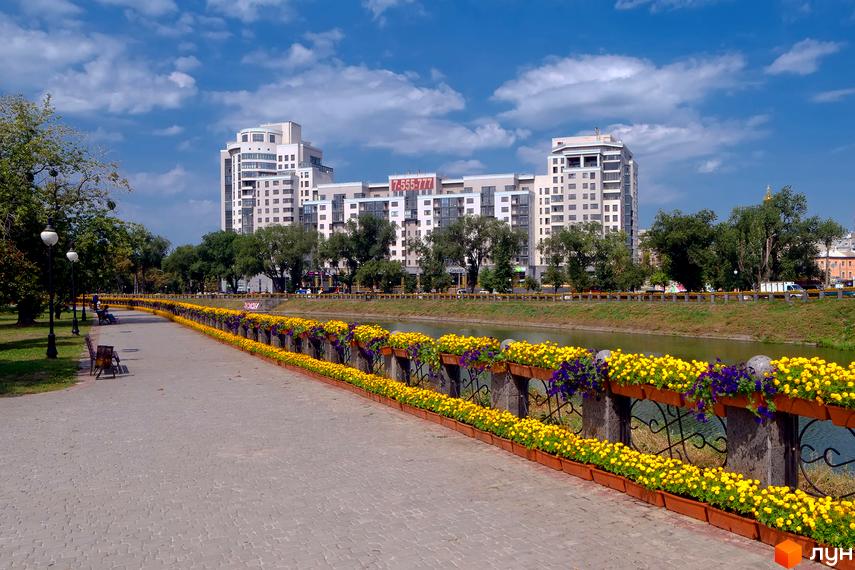 Будинок на Набережній: сучасний житловий комплекс з квітковими клумбами вздовж набережної та річкою.