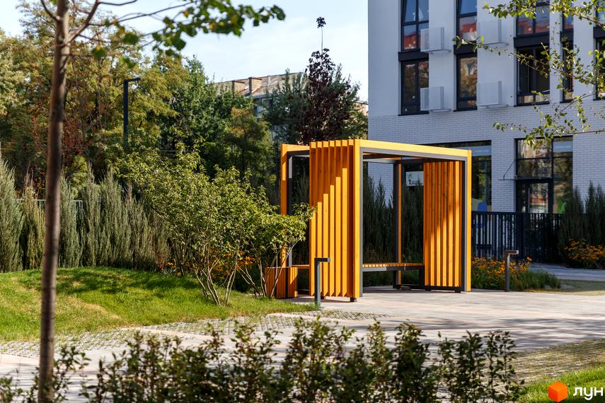 Житловий комплекс Файна Таун: білий багатоповерховий будинок з темними вікнами, поруч зелений газон та жовта дерев'яна пергола.