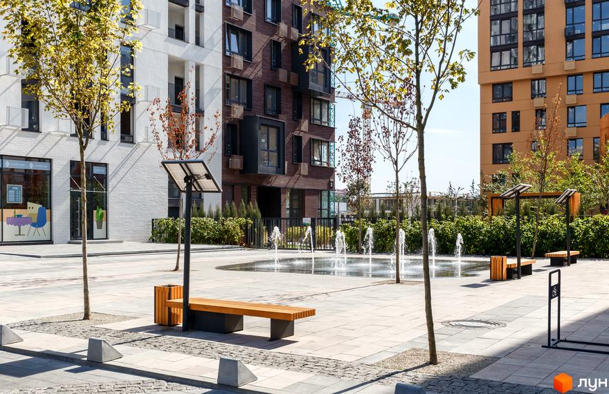 Зовнішній вигляд будинків ЖК Файна Таун. На території є лавки, дерева та фонтан. На передньому плані бруківка.