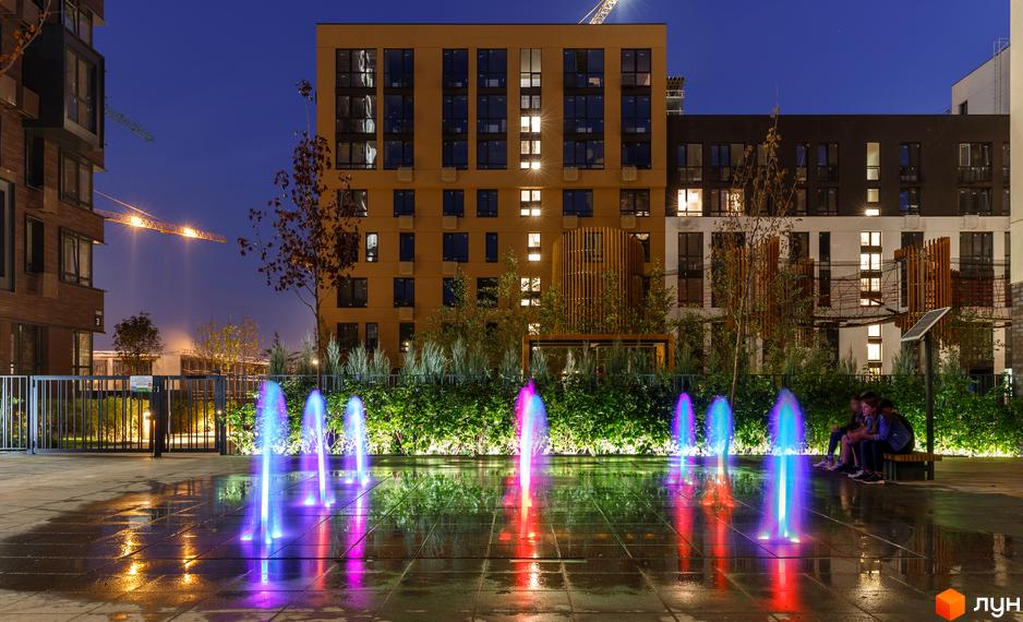 Житловий комплекс Файна Таун з підсвіченими фонтанами та лавками для відпочинку. На задньому плані — будівлі з великими вікнами, що освітлюються ввечері.