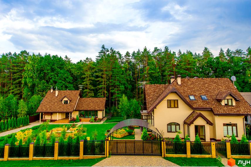 Двоповерховий будинок з коричневою черепицею на даху. Перед будинком газон, ковані ворота та доглянута прибудинкова територія.