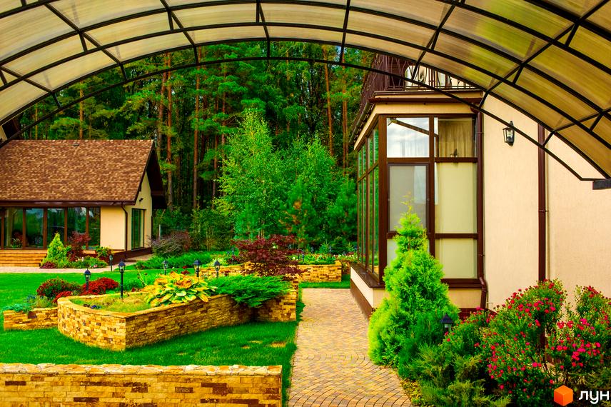 Затишний будинок з коричневою черепицею на даху та великими вікнами. Доглянута прибудинкова територія з газоном, доріжками та декоративними рослинами в Котеджному містечку Лелечий Хутір.