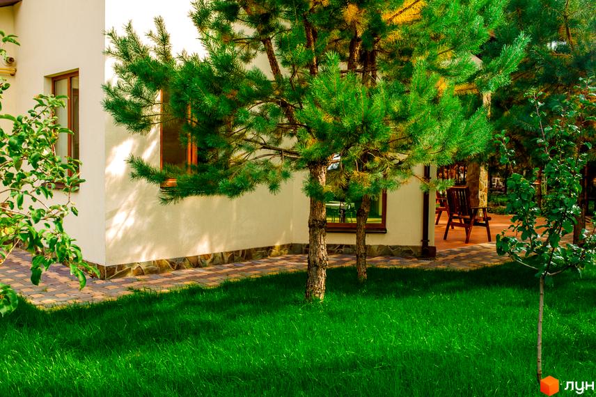 Білий будинок з вікнами та зеленими деревами на подвір'ї. Поруч доглянутий газон та бруківка. КМ Лелечий Хутір - ідеальне місце для життя.