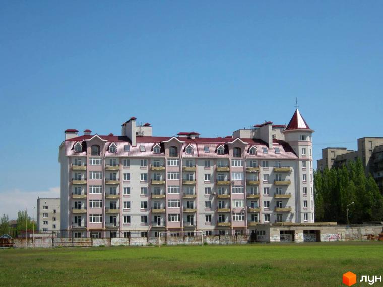 Зовнішній вигляд багатоповерхового будинку з червоним дахом та балконами на фоні блакитного неба. На передньому плані зелений газон.