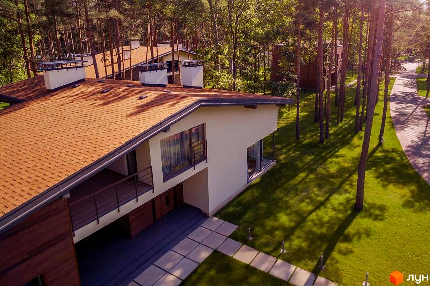 Житловий комплекс Goodlife Park з коричневим дахом та великими вікнами. Навколо будинку зелений газон та сосновий ліс.