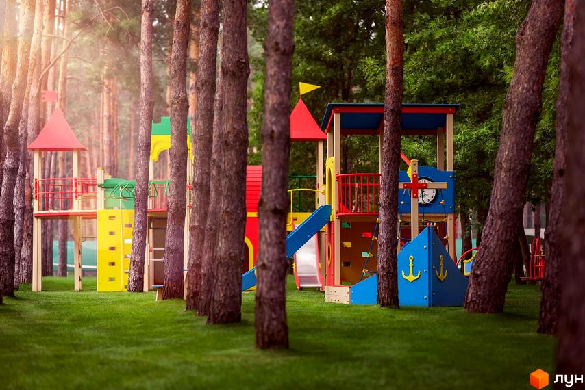 Котеджне містечко Goodlife Park з дитячим майданчиком серед сосен. Яскраві гірки, драбинки та вежі для ігор на зеленій галявині.