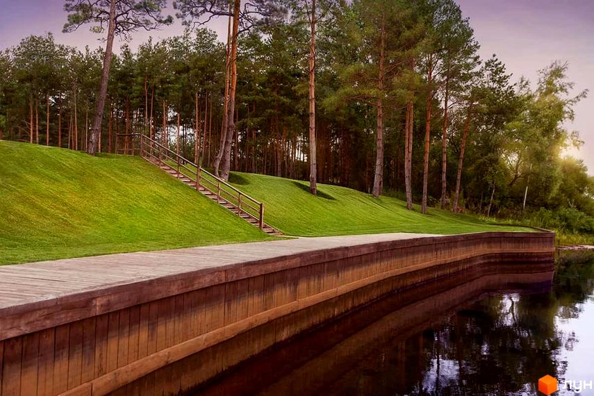 Зображення прибережної зони Котеджного містечка Goodlife Park. Вид на дерев'яний пірс та зелений схил з дерев'яними сходами до лісу.
