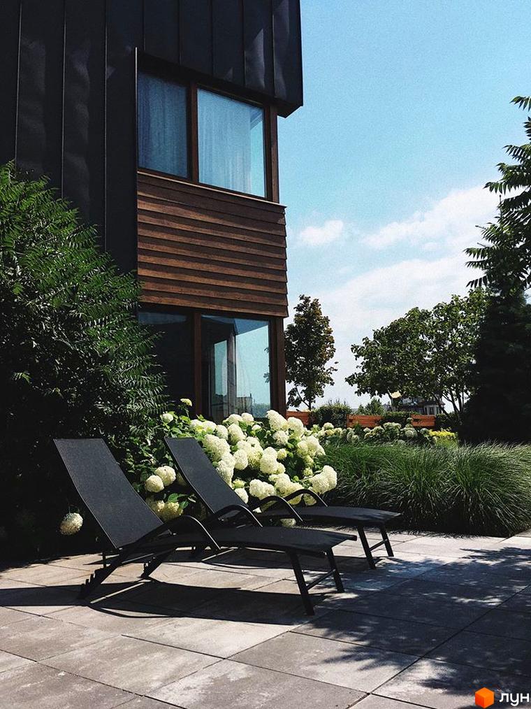 Терасований будинок з темним фасадом. Біля будинку розташовані шезлонги, квіти та газон, створюючи атмосферу відпочинку.