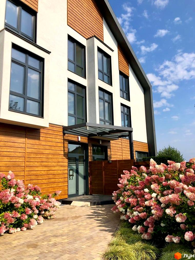 Зовнішній вигляд будинку з дерев'яним оздобленням фасаду та великими вікнами. На передньому плані квітучі кущі та бруківка.