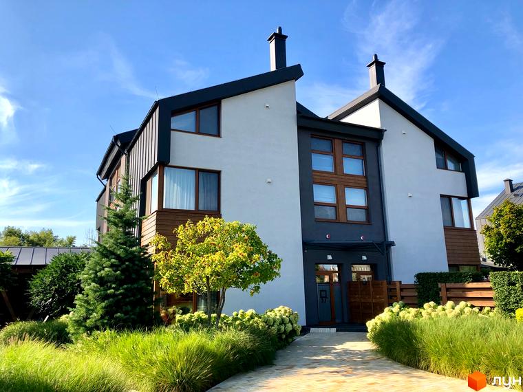 Двоповерховий будинок з білими стінами та темною покрівлею. На прибудинковій території газон, дерева та доріжка з бруківки.