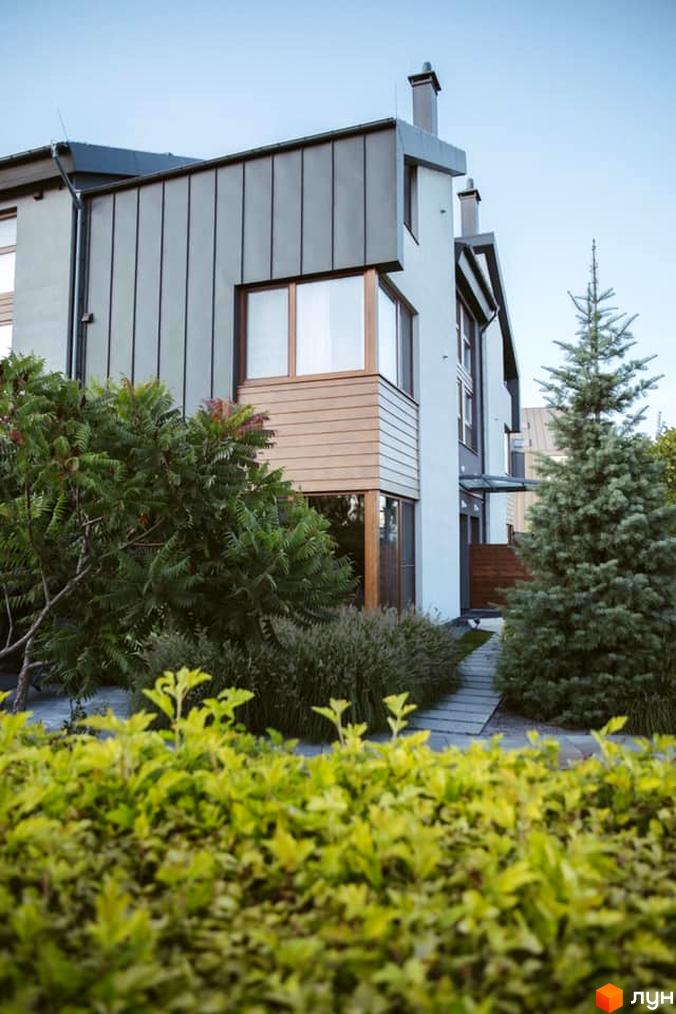 Зображення фасаду будинку з сірим металевим дахом та великими вікнами. На передньому плані зелені кущі та доріжка, що веде до входу.