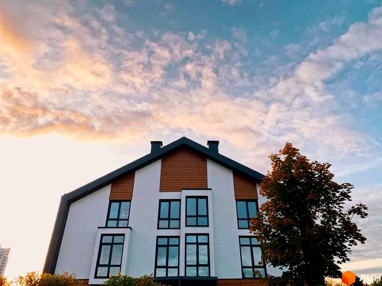 Зображення сторінки житлового комплексу. На фото зображений білий будинок з великими вікнами та дерев'яними вставками. Навколо будинку ростуть дерева, а на фоні небо з хмарами.