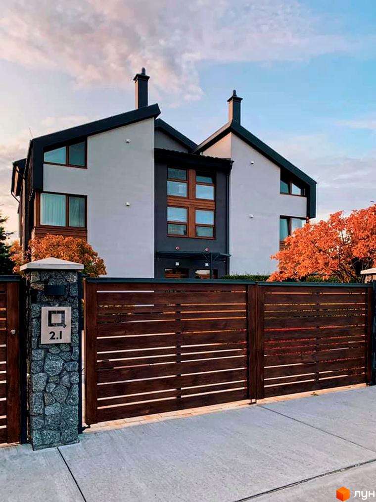 Двоповерховий будинок з темним дахом та дерев'яним парканом. На території є зелені насадження та осінні дерева, що додають затишку.