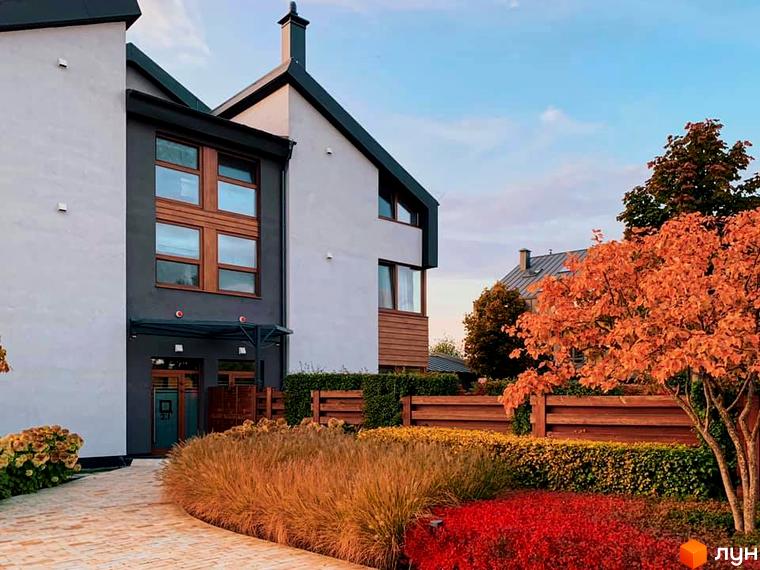 Терасований будинок з великими вікнами та вхідною групою. На території розташовані доглянуті газони з рослинами та дерева.