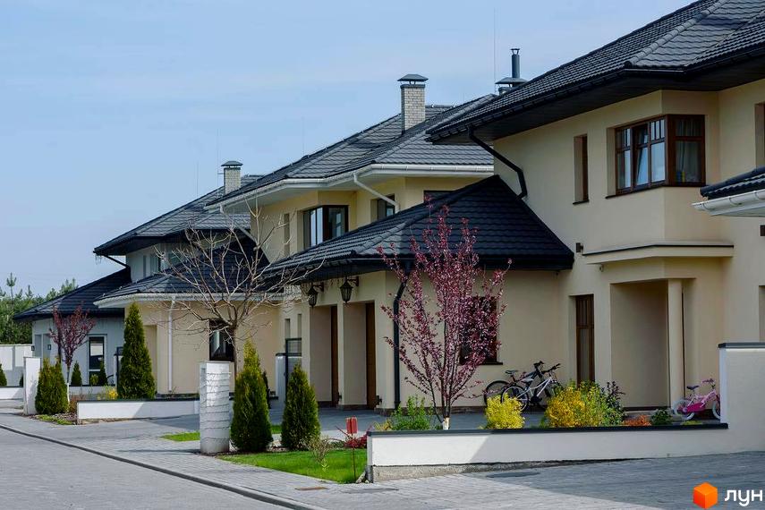 Котеджне містечко КМ Belgravia: фасади будинків світло-бежевого кольору з темними дахами, акуратні зелені насадження та доріжки. На території є дитячі велосипеди.
