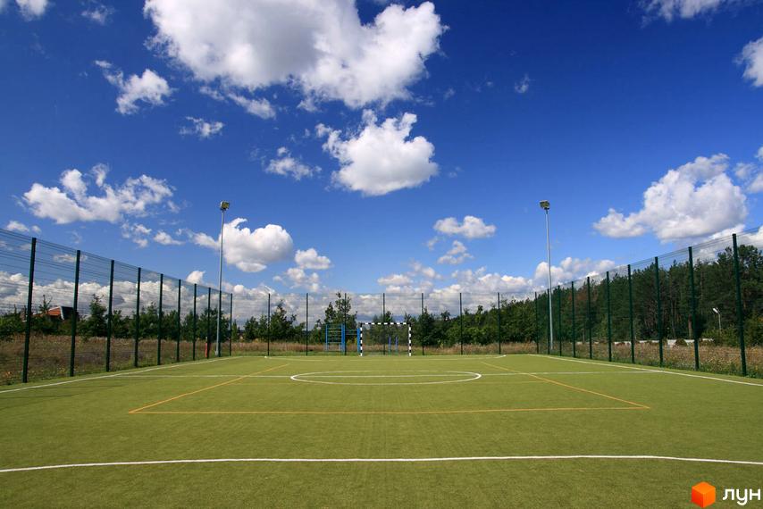 Футбольне поле в Котеджному містечку КМ Бобриця-Парк. Зелене поле з розміткою, огорожене сіткою, на тлі блакитного неба з хмарами.