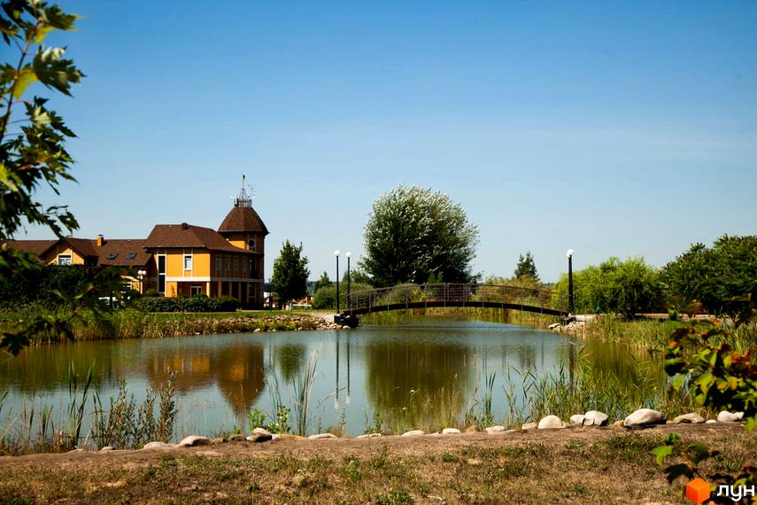 Житловий комплекс КМ Бобриця-Парк з жовтим будинком з баштою, що стоїть біля водойми з містком. Навколо будинку зелені дерева та кущі.