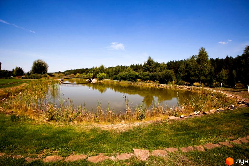 Озеро з містком в Котеджному містечку Бобриця-Парк. Навколо озера зелена трава та дерева, що створюють мальовничий пейзаж.