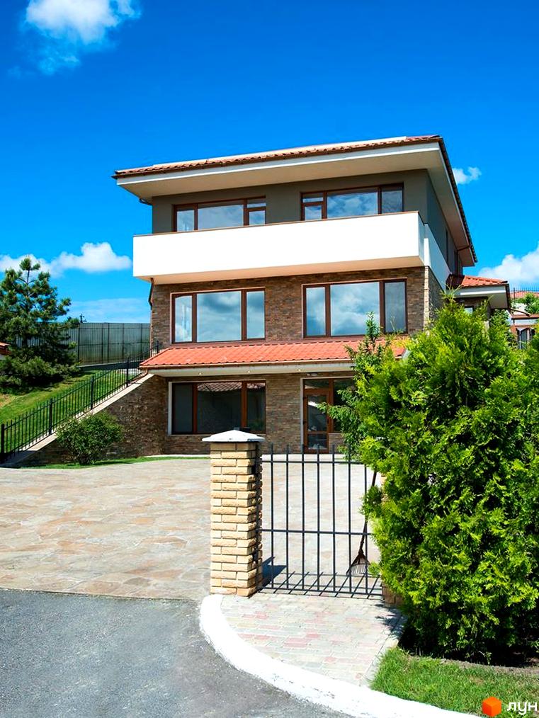 Триповерховий будинок з великими вікнами та терасою. Перед будинком розташована доглянута прибудинкова територія з рослинами та кованим парканом.