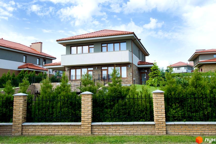 Двоповерховий будинок з терасою та панорамними вікнами. Перед будинком розташована зелена зона з туями та цегляним парканом.