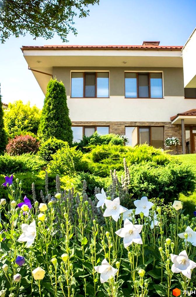 Фасад будинку з великими вікнами та терасою. Перед будинком розташована доглянута прибудинкова територія з квітами та зеленню.