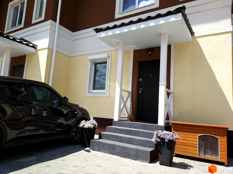 Таунхауси „Плюс“: двоповерховий будинок з коричневим дахом та світло-жовтими стінами, білим ганком та чорними дверима. Перед будинком припаркований чорний автомобіль та стоять дві вазони з квітами.