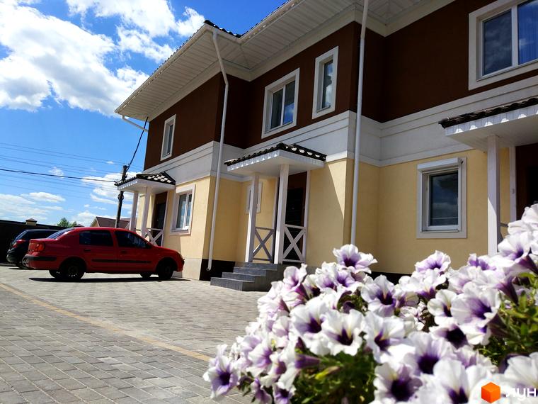 Двоповерхові таунхауси „Плюс“ з коричнево-жовтим фасадом та білими елементами. Перед будинками розташована вимощена парковка з червоним автомобілем та квітами.