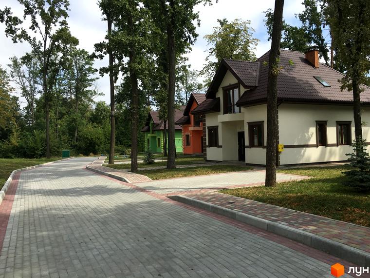 Котеджне містечко Козак хутір: затишні будинки з коричневими дахами та світлими фасадами, розташовані вздовж вимощеної бруківкою дороги з зеленими насадженнями.