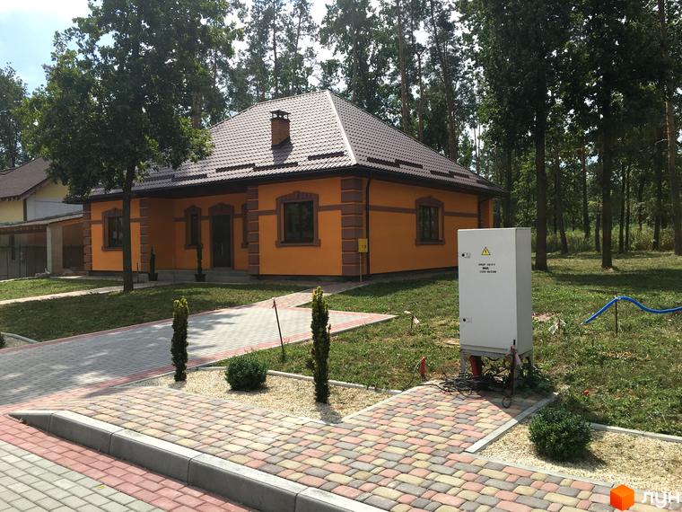 Котеджне містечко Козак хутір: одноповерховий будинок з помаранчевими стінами та коричневим дахом, розташований серед дерев. Прибудинкова територія з бруківкою та зеленими насадженнями.