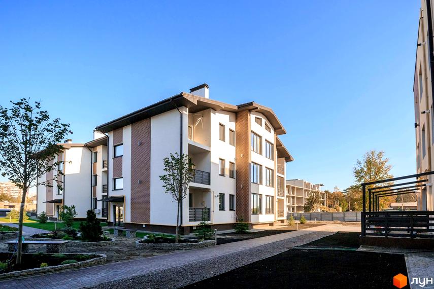 Житловий комплекс Клеменова Дача з білими фасадами та коричневою цегляною обробкою, темним дахом. Навколо будинку доріжки, зелені газони та молоді дерева.