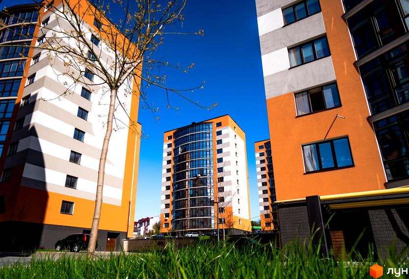 Зовнішній вигляд багатоповерхового будинку в Житловому комплексі Містечко Соборне на фоні блакитного неба. Вид на прибудинкову територію з травою.