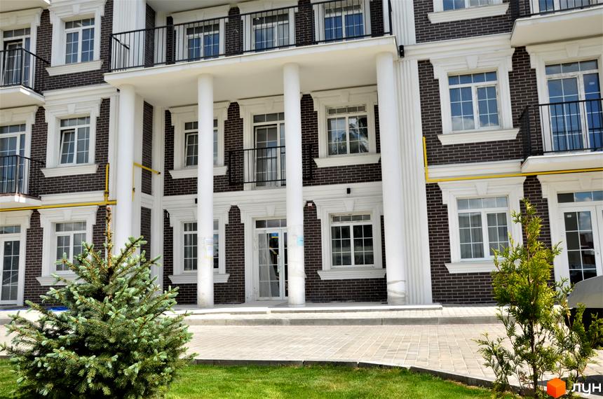 Житловий комплекс Britania Hall з темною цегляною фасадою, білими колонами та балконами. Навколо будинку викладена бруківка та зелений газон.