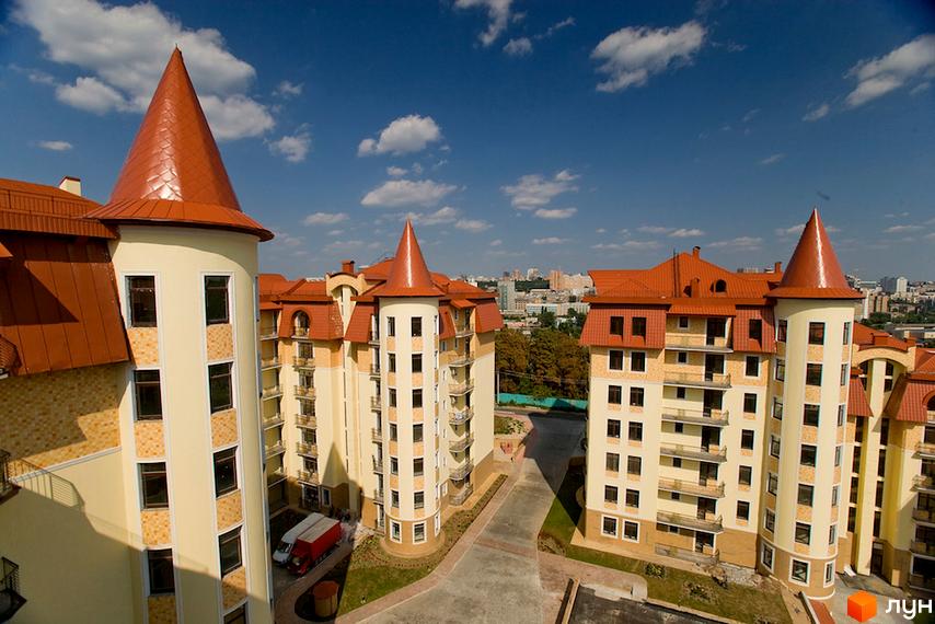 Житловий комплекс Альпійське містечко: будинки світлих тонів з помаранчевими дахами-вежами та балконами. Навколо облаштована територія з доріжками та зеленню.