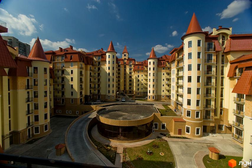 Житловий комплекс Альпійське містечко: будівлі світлого кольору з червоними дахами та вежами. На території комплексу є доріжки, зелені газони та в'їзд.