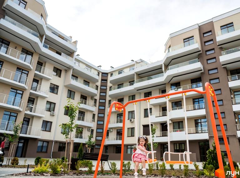 Житловий комплекс GREEN LAND з дитячим майданчиком та дівчинкою на гойдалці. Сучасна архітектура новобудови з балконами та вікнами.