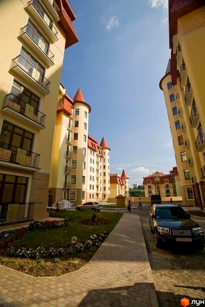 Житловий комплекс Альпійське містечко: будинки світлого кольору з червоними дахами та вежами. Прибудинкова територія з доріжками, газонами та клумбами.