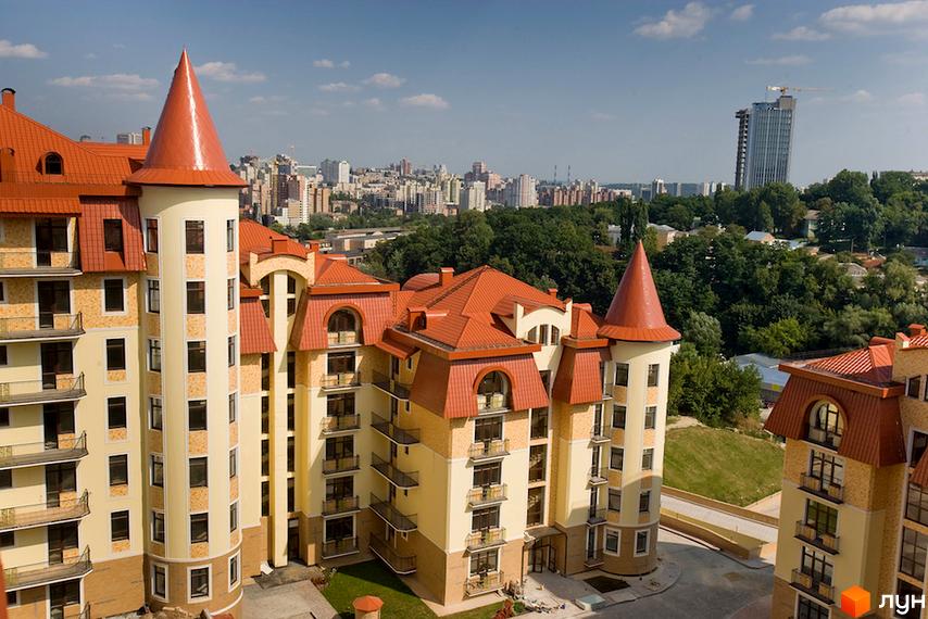 Житловий комплекс Альпійське містечко: будинки світлого кольору з червоними дахами та конічними вежами, оточені зеленою територією та міським пейзажем.