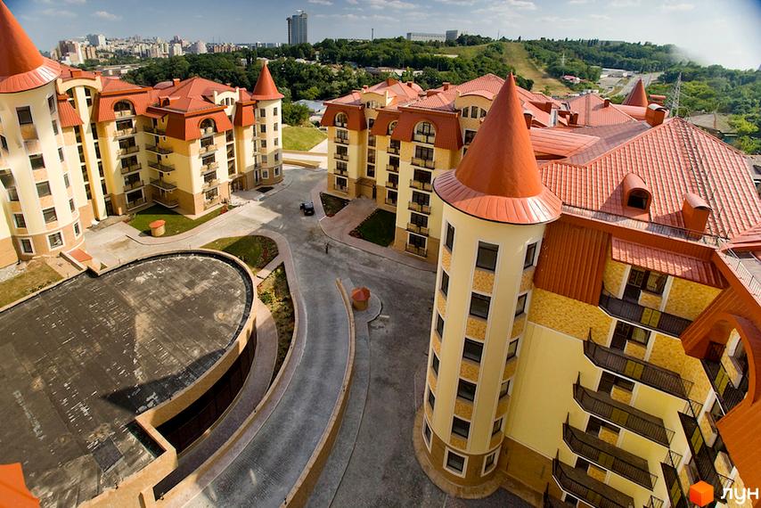 Житловий комплекс Альпійське містечко: будинки світло-жовтого кольору з помаранчевими дахами та вежами, оточені дорогами з бруківки та зеленими газонами.