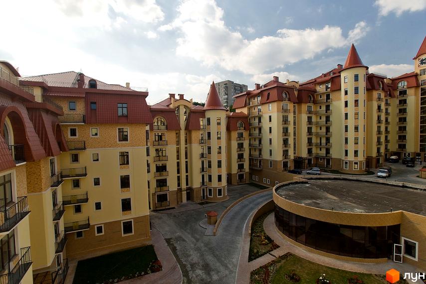 Житловий комплекс Альпійське містечко з бежевими фасадами, червоними дахами та вежами. Навколо комплексу видно доріжки та зелені насадження.