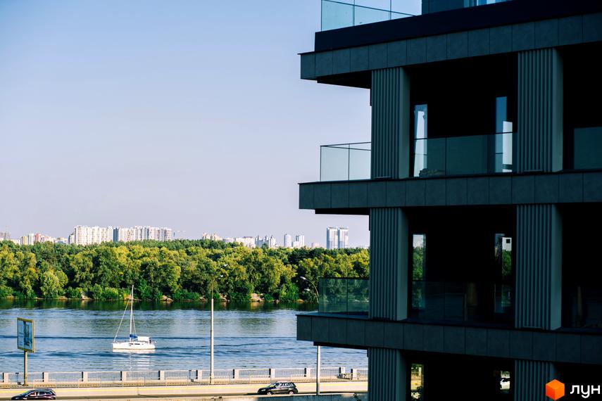 Фасад будинку Житловий район Rybalsky з темними панелями та скляними балконами. На передньому плані видно річку з човном, міст та зелений берег з міськими забудовами.