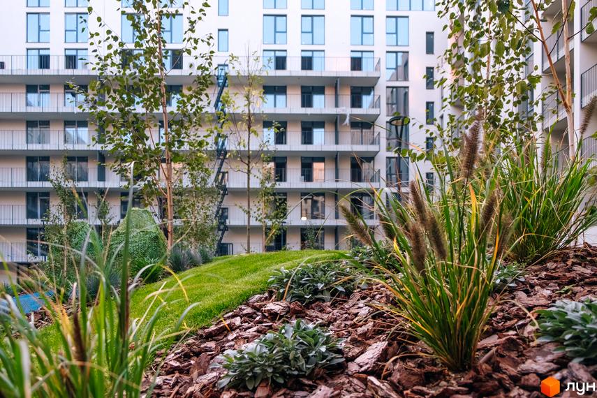 Фасад багатоповерхового будинку Rybalsky з балконами та вікнами. Прибудинкова територія з газоном, деревами та декоративними рослинами.