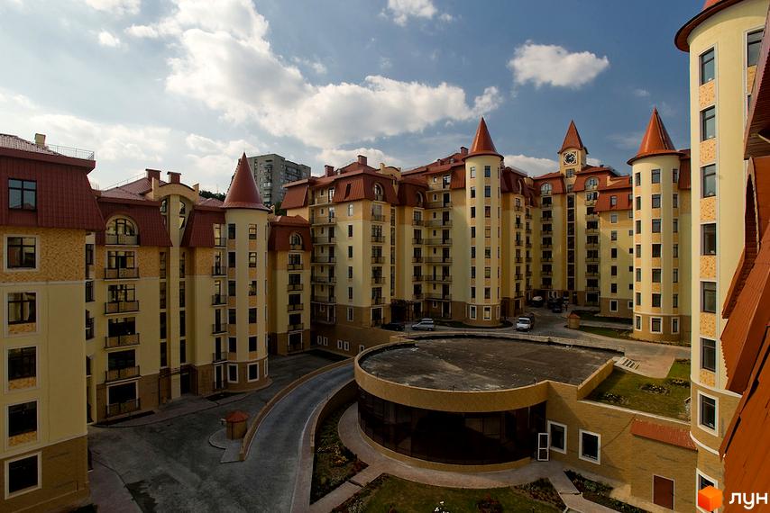 Житловий комплекс Альпійське містечко: світло-жовті багатоповерхові будинки з червоними дахами та вежами. У дворі вимощена дорога, зелені насадження та кругла будівля.