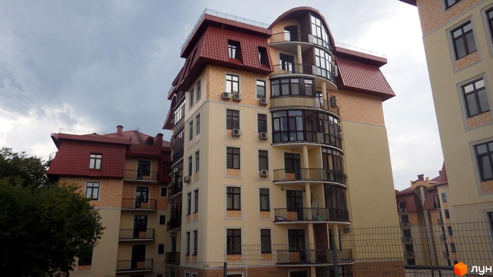 Житловий комплекс Альпійське містечко з бежевими фасадами, червоними дахами та скляними балконами. На передньому плані огорожа та дерева.