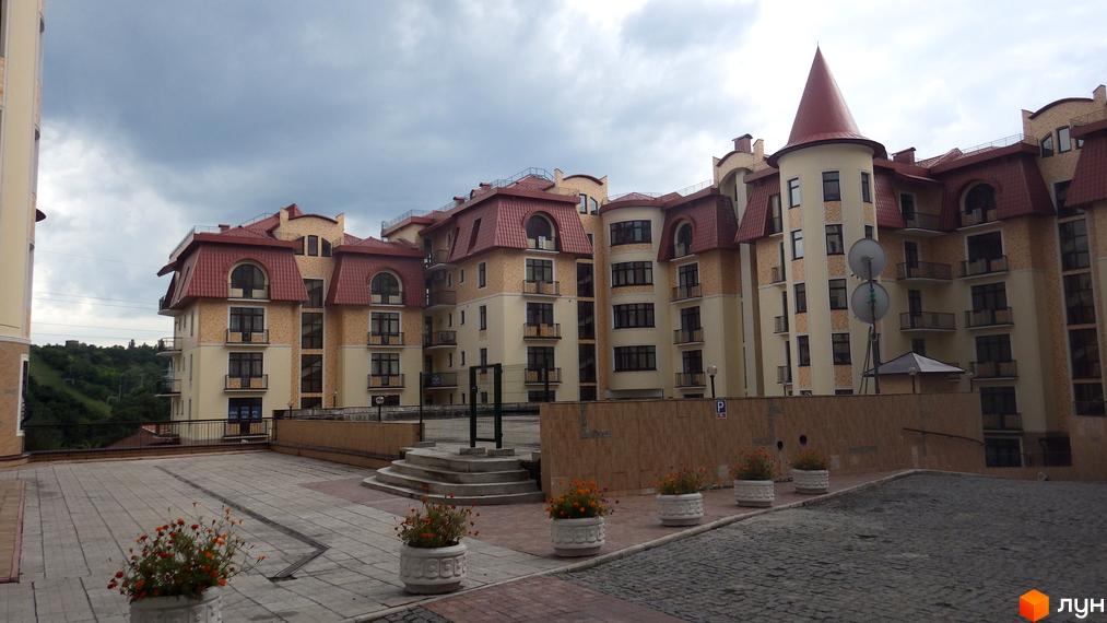 Житловий комплекс Альпійське містечко: будівлі зі світлими фасадами, червоними дахами та вежею. На передньому плані вимощений двір з клумбами.