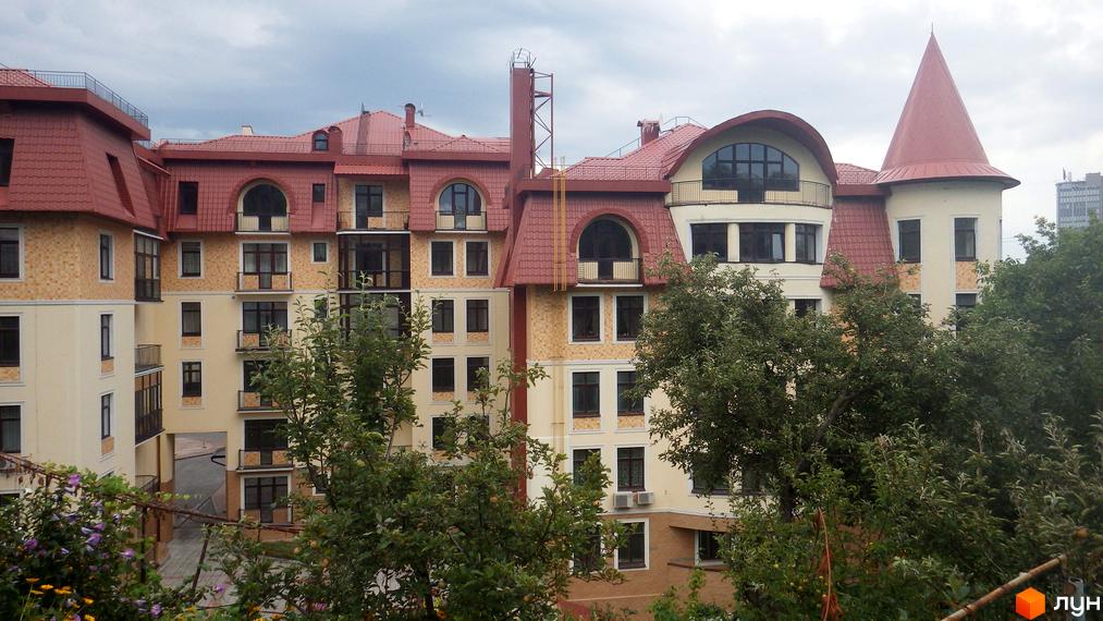 Житловий комплекс Альпійське містечко: будівля з червоним черепичним дахом, світлими фасадами, арковими вікнами та конусоподібною вежею, оточена зеленими деревами.