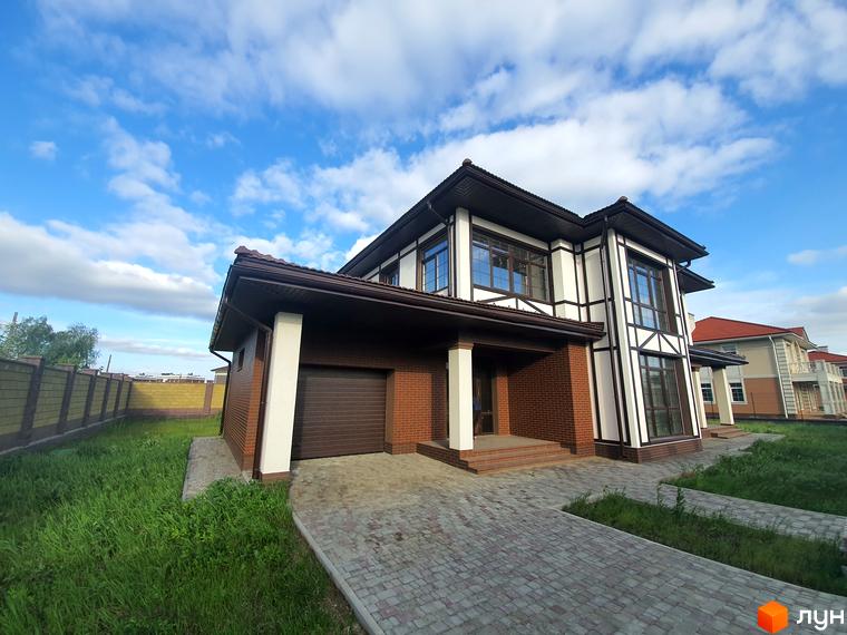 Двоповерховий будинок з коричневою черепичною покрівлею та білими колонами. Перед будинком викладена бруківка, газон з зеленою травою.