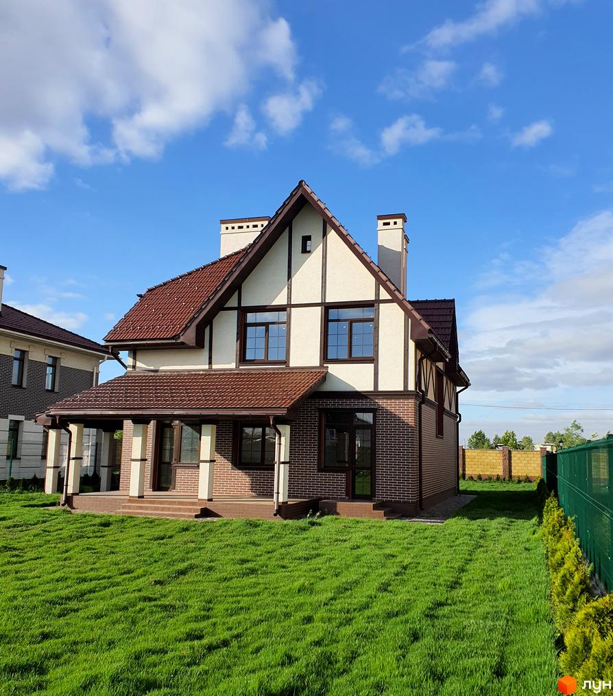 Двоповерховий будинок з коричневою черепичною покрівлею та цегляною кладкою. На подвір'ї зелена трава та доглянута прибудинкова територія.