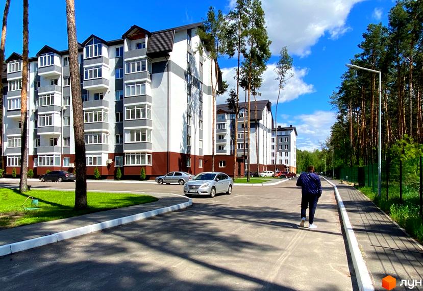 Житловий комплекс Ідея з багатоповерховими будинками біло-сіро-коричневого кольору, оточений зеленими газонами та високими соснами. Поруч дорога та ліс.